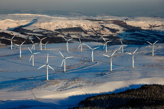 Snow-covered landscape at sunrise with a wind farm of multiple turbines standing tall against a hilly backdrop.