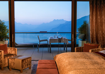 Elegant hotel room with a view of a mountain range during twilight, featuring a plush bed, seating area, and a dining setup on the balcony.