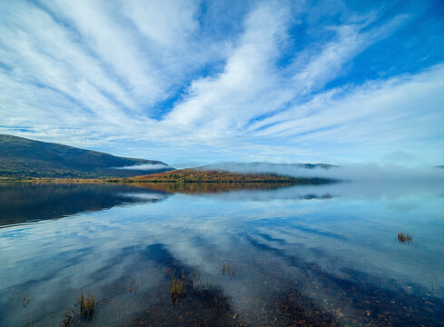 Serene lake reflecting a blue sky with streaks of clouds, surrounded by hills and a thin layer of mist above the water. Loch Lochy