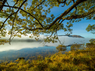Mist hovers over a serene lake with clear reflections of trees and a mountain under a blue sky. Loch Arkaig
