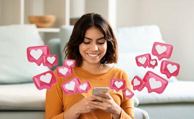 Arabic young woman with short brown hair is sitting in a living room, smiling as she uses her smartphone. Pink icons with white hearts inside are floating around her, appearing to be notifications.