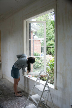 A woman in casual attire is renovating a house, scraping paint from a window frame in a room under construction, New Zealand