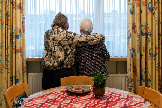 Two elderly women embrace while looking out the window, a cozy room with a flowered curtain and a fruit bowl on the table suggests a homely atmosphere.