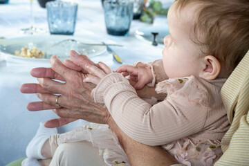 An infant in a pink outfit is held in the arms of a senior, whose wrinkled hands gently grasp the baby's tiny ones at a dining table.