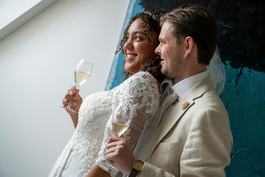 A bride in a lace dress and a groom in a suit are smiling and toasting with white wine in front of an abstract painting.