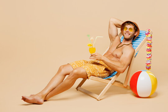 Full body young sexy man wear yellow shorts swimsuit hat relax rest near hotel pool sit in deckchair drink orange cocktail isolated on plain beige background. Summer hot vacation sea sun tan concept.