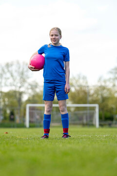 Fototapeta A smiling young girl in blue sportswear holds a pink soccer ball on a grassy field.