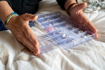 Close-up of a person organizing a weekly pill organizer with various medications on a white bedspread.
