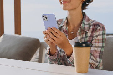 Close up cropped young happy woman wear shirt casual clothes sitting alone at street cafe table in coffee shop drink tea hold in hand use mobile cell phone rest in restaurant during free time outdoor.