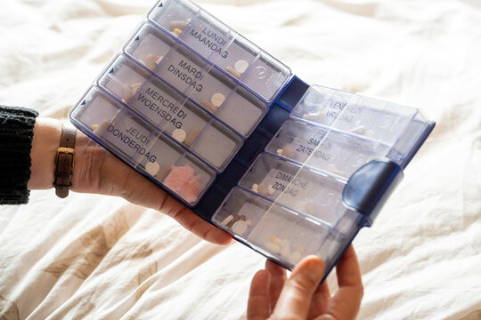 Hand holding a pill organizer with compartments labeled with days of the week in different languages, filled with various pills.