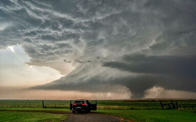 A storm chaser's vehicle parked on a dirt road observing a massive tornado forming under a dark, dramatic sky in an open field.