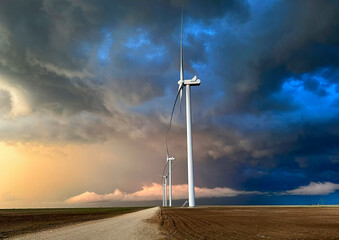 Sunset sky with storm clouds looming over a wind farm featuring two prominently displayed wind turbines standing on an open field.