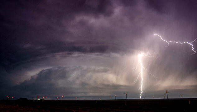 A dramatic nocturnal landscape showcasing a brilliant lightning strike illuminating wind turbines under a stormy sky.