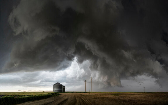 Ominous storm clouds loom over a rural road with a solitary building and a telephone pole, suggesting an impending severe weather event.