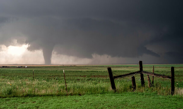 A tornado touches down in a rural landscape near a wooden fence under a dramatic stormy sky.