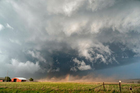 A tornado forms under a menacing thunderstorm over a rural landscape with a red barn and a wooden fence in the foreground.