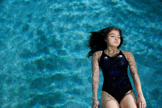 A young girl floats serenely in a clear blue swimming pool wearing a blue and black swimsuit.