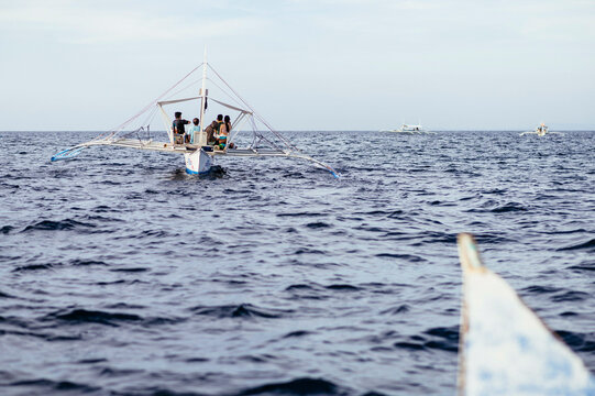 Traditional outrigger canoe sailing across the open sea with people onboard and other boats visible in the distance.
