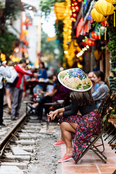 A woman wearing a traditional conical hat sits on a chair by a bustling street adorned with lanterns.