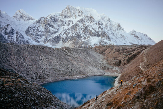A serene mountain landscape with a clear blue alpine lake and two hikers on a trail, with rugged snowy peaks in the background.