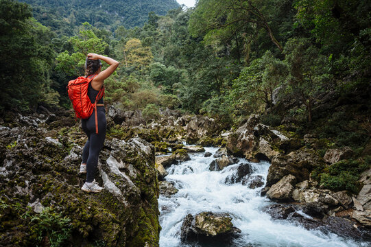 A hiker with a red backpack stands on a rocky outcrop overlooking a rushing river in a lush forest.