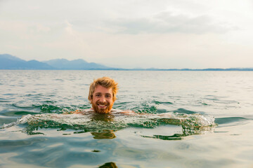 Smiling man swimming in a serene lake with mountains in the distance at sunset.