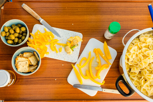 A colorful cooking preparation scene with chopped yellow bell peppers, olives, crackers, and uncooked pasta on a wooden surface.