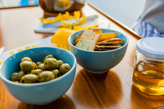 A variety of snacks arranged on a wooden table with crackers, cheese, green olives, and honey in a jar.