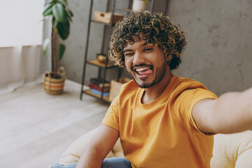 Close up young happy fun Indian man wear orange casual clothes do selfie shot pov on mobile cell phone sit in armchair stay at home hotel flat rest relax spend free time in living room Lounge concept