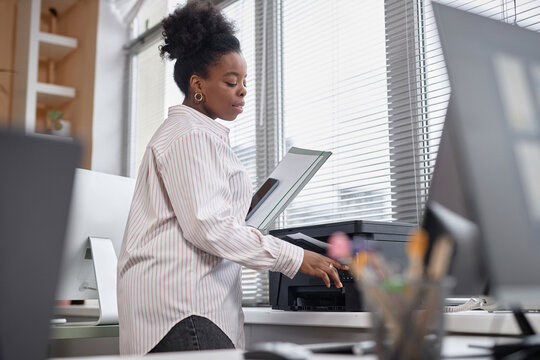 Medium shot of young female African American employee or student in casual white shirt using printer in office or class, copy space