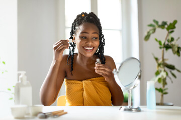 African American young woman with long black braids applies facial serum to her skin, looking in a...