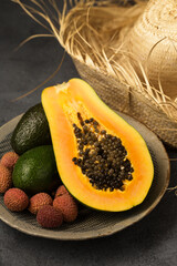 Two avocados, half a papaya and lychee berries on a gray clay plate near the straw hat