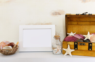 Image of old chest full of seashells and starfish next to empty photo frame over white shelf and pastel wooden background. For mockup