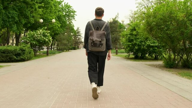 A young guy with a backpack on his back walks through the spring city. Back view.
