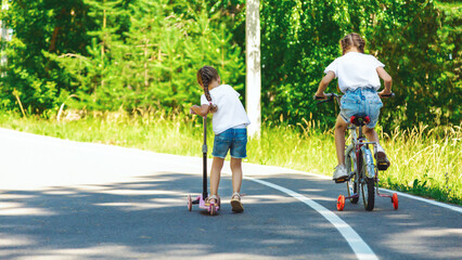 little girls riding on bicycle and scooter on road in the park on summer day