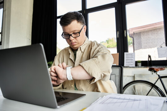 A young man with Down syndrome sits at a desk with a laptop open, checking his watch.