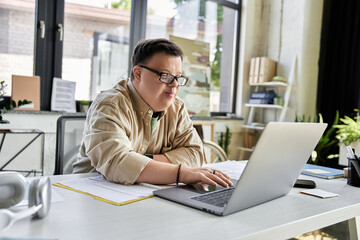 A young man with Down syndrome sits at a desk in an office, concentrating on his laptop.