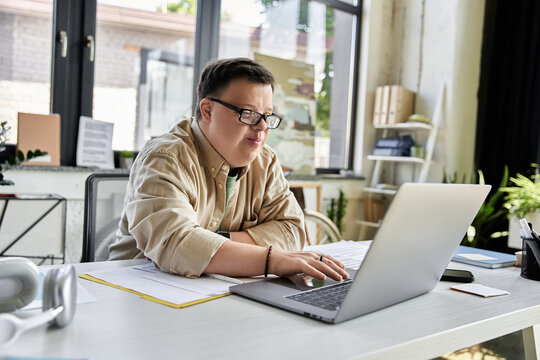 A young man with Down syndrome sits at a desk in an office, concentrating on his laptop.