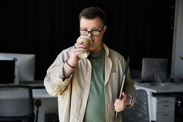 Young man with Down syndrome sips coffee while holding a laptop.