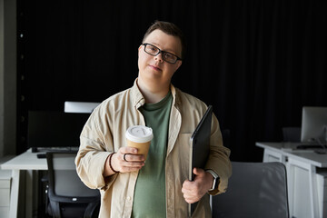 Young man with Down syndrome wears glasses, holding coffee and laptop in office