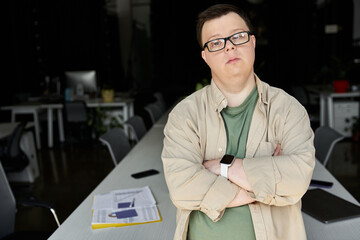 A man with Down syndrome stands confidently in an office setting with arms crossed.