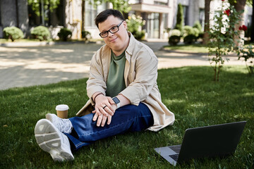 A young man with Down syndrome sits on the grass, enjoying a coffee and his laptop.