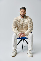 A bearded man in a white sweater striking a pose while sitting on a blue chair in a studio with a...