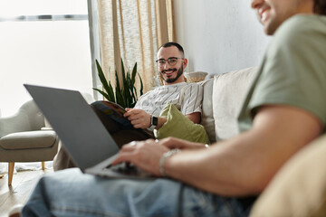 Fototapeta premium Couple relaxes on couch, one works on laptop while other reads magazine, sharing glance