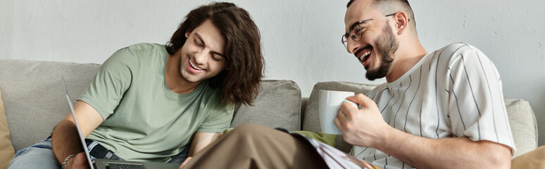 Obraz premium A gay couple on a couch, one looking at a laptop while the other laughs and holds a mug.