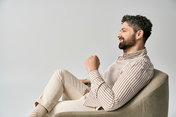 A stylish bearded man in elegant attire sits on armchair against a grey studio background.