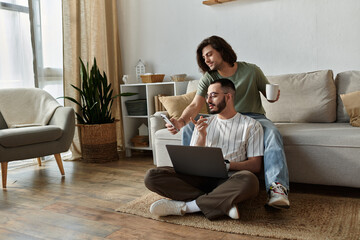 A couple sits on the floor, one man using a laptop while the other looks at his phone.