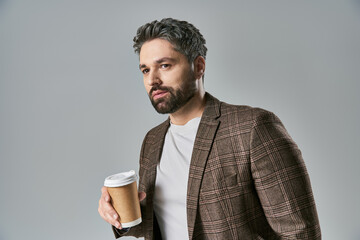 A stylish man with a beard elegantly holds a cup of coffee, exuding charm and sophistication against a grey backdrop.