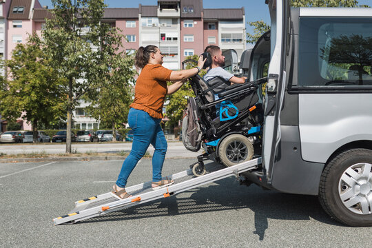 Female assistant helping a man with disability to enter an accessible vehicle, pushing him over a wheelchair car ramp.
