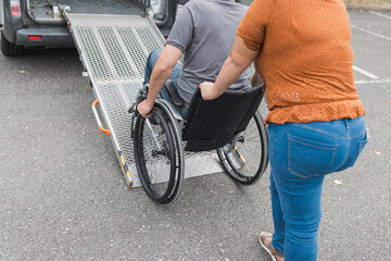 Female assistant helping a male person in wheelchair with transport using accessible vehicle van ramp. Disability and mobility concepts.